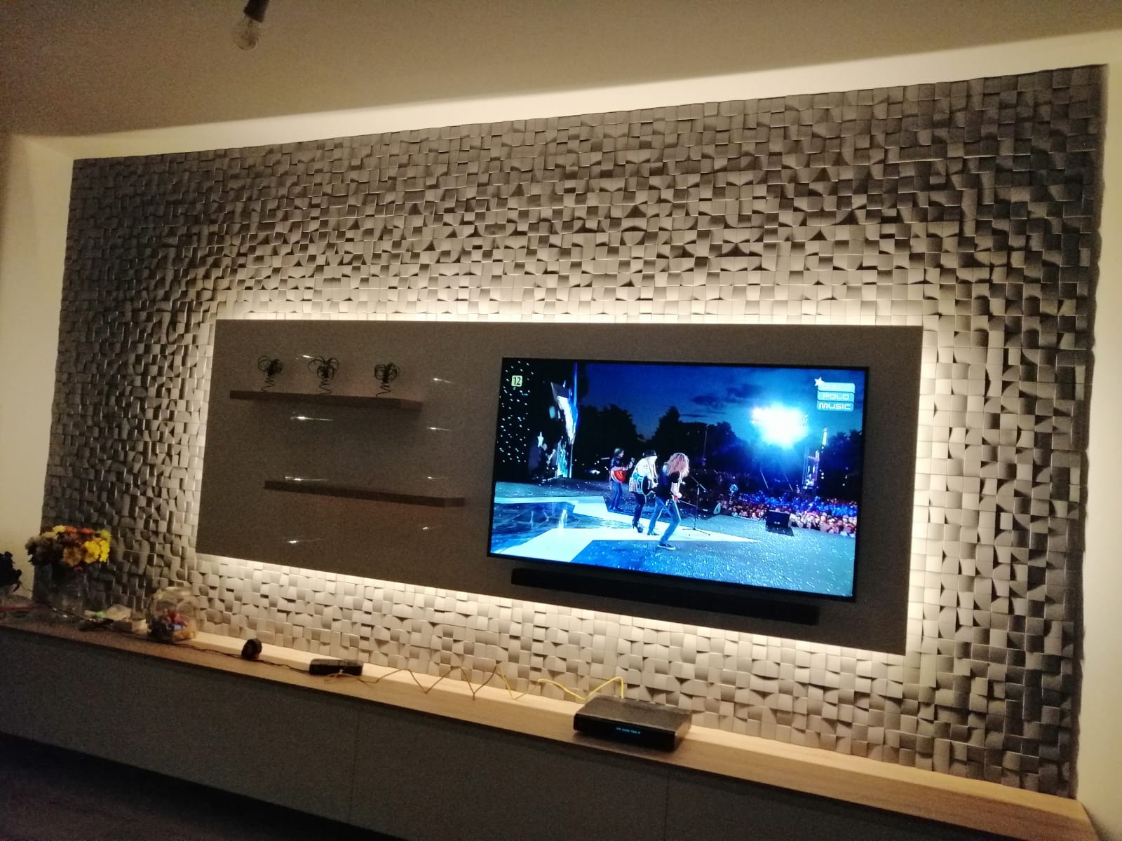 Ściana telewizyjna z trójwymiarową, podświetloną strukturą paneli dekoracyjnych, z półkami i telewizorem wyświetlającym koncert.