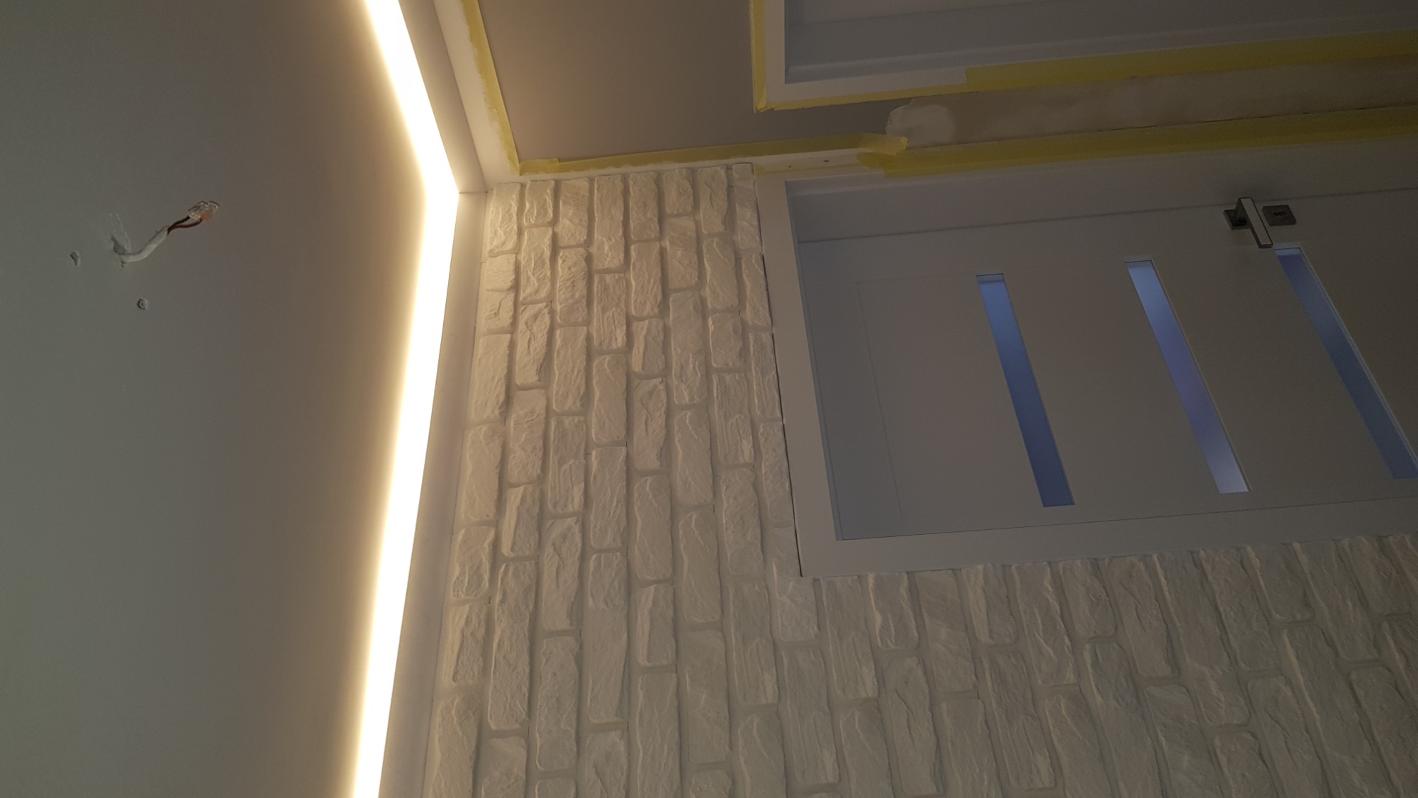 Ściana wykończona białą cegłą dekoracyjną, z widoczną białą stolarką drzwiową z przeszkleniami i oświetleniem LED wzdłuż sufitu, widoczne przewody elektryczne na ścianie.