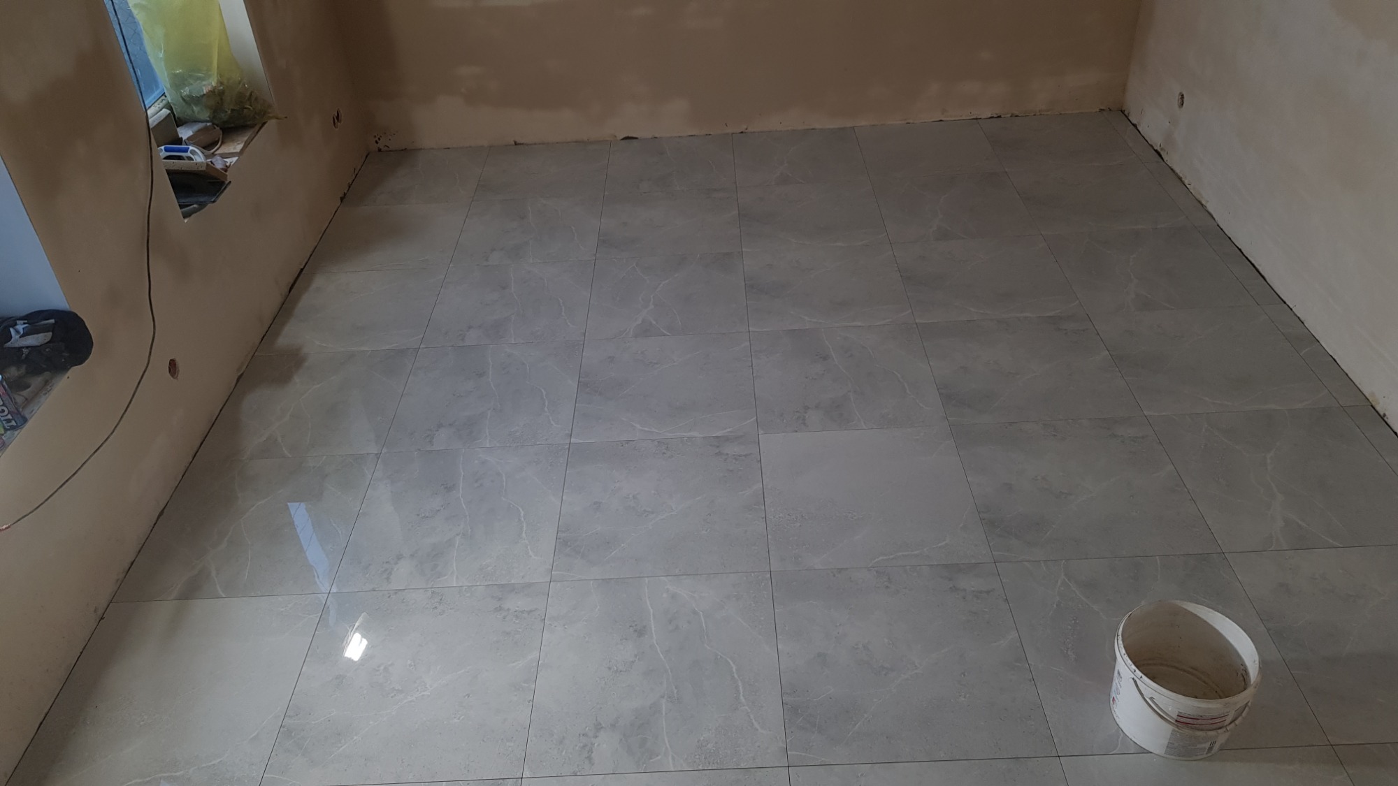 Ułożona szara, marmurkowa podłoga z płytek w trakcie prac wykończeniowych. Widoczny biały kubeł na farbę w rogu pomieszczenia i niedokończone ściany.