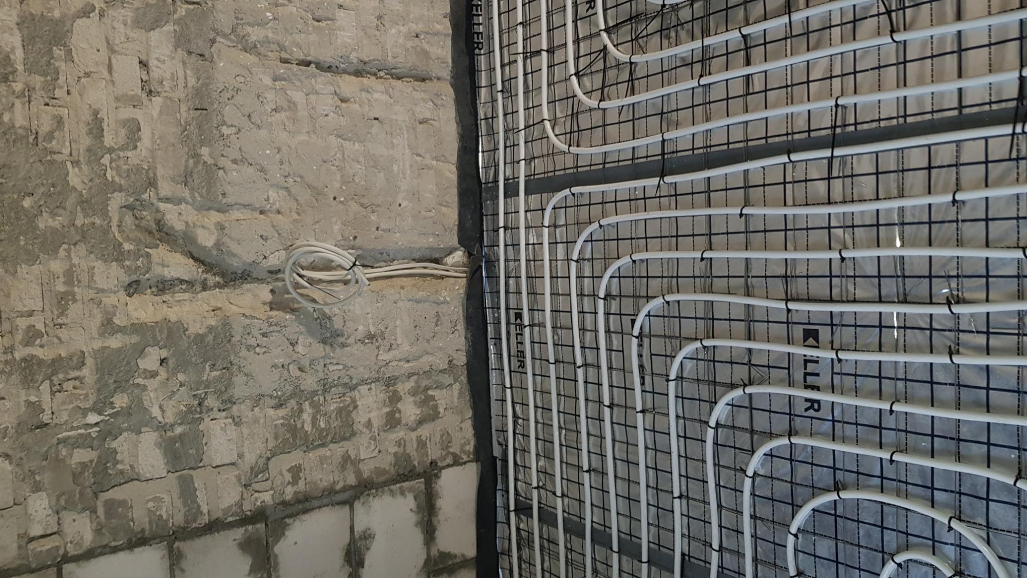 Montaż ogrzewania podłogowego na ścianie: białe rurki układane na siatce montażowej, surowa ściana z cegły w tle.