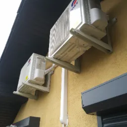 Dwa klimatyzatory marki Gree zamontowane na elewacji budynku o żółtej strukturze, widoczne wsporniki montażowe i rura odprowadzająca skropliny, drabina oparta o ścianę.