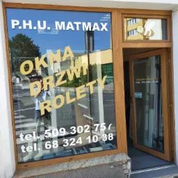 Fasada firmy P.H.U. MATMAX z otwartymi drzwiami, oferującej okna, drzwi i rolety, z widocznym odbiciem osoby robiącej zdjęcie w szybie wystawowej.