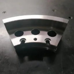 Metalowy element w kształcie wycinka koła z trzema okrągłymi otworami i dwoma wycięciami, leżący na ciemnej, matowej powierzchni.