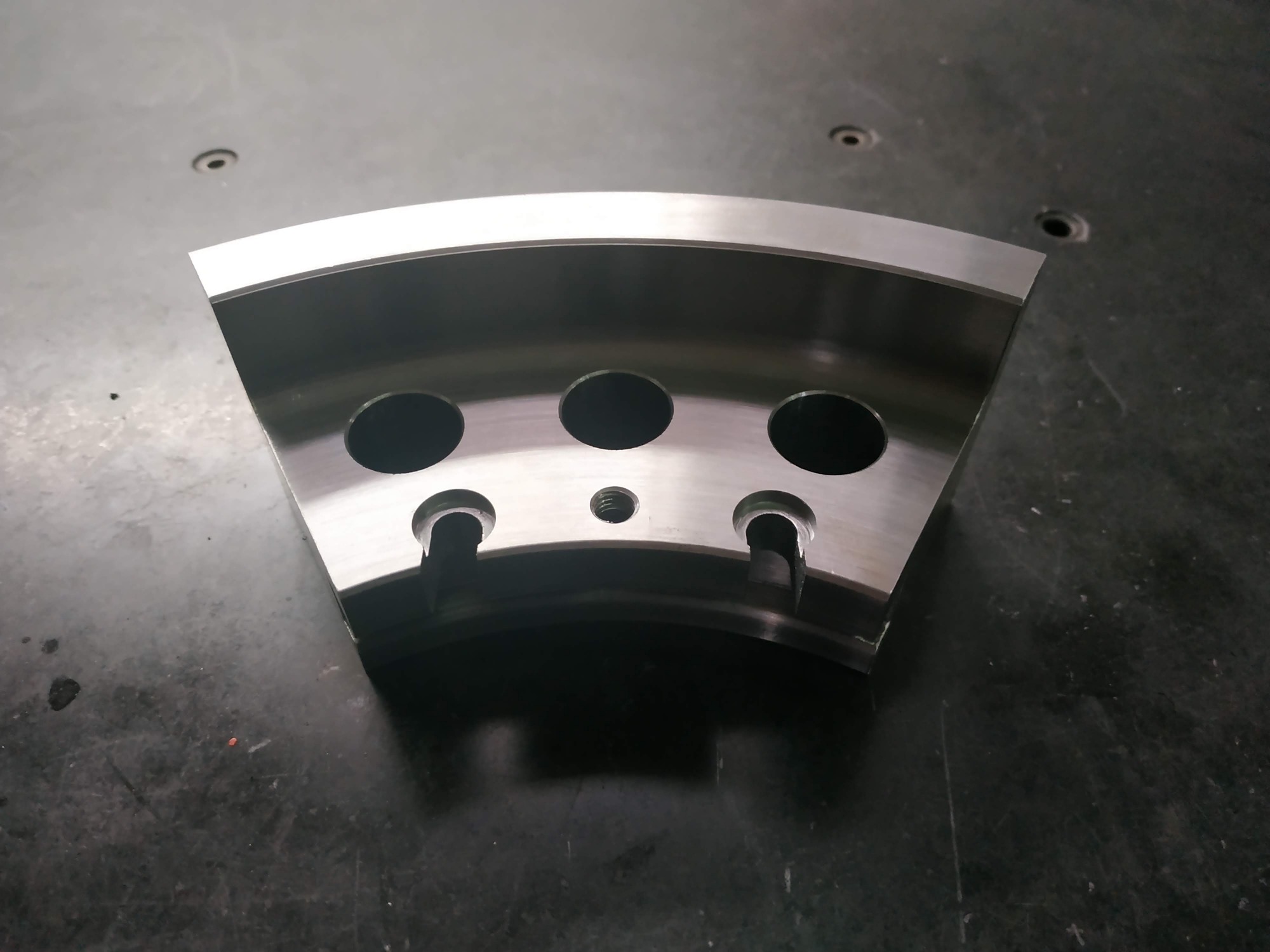 Metalowy element w kształcie wycinka koła z trzema okrągłymi otworami i dwoma wycięciami, leżący na ciemnej, matowej powierzchni.