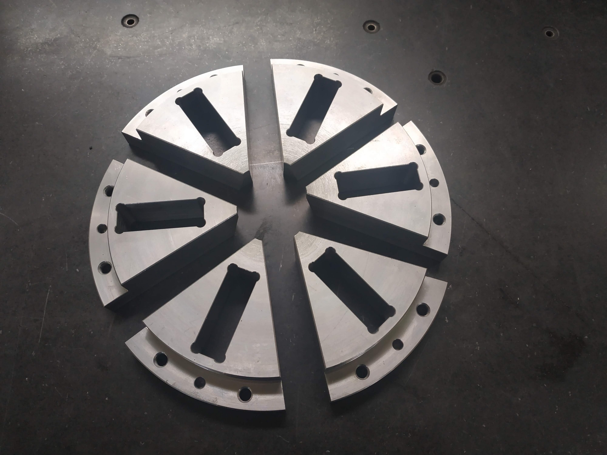 Sześć segmentów metalowej tarczy z otworami i prostokątnymi wycięciami, leżących na szarej powierzchni, tworzących okrąg.