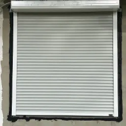 Zamknięta, jasnoszara roleta zewnętrzna zamontowana w oknie budynku z pustaków, widoczne uszczelnienie ramy okiennej czarną masą. Nad roletą widoczne logo producenta na osłonie.