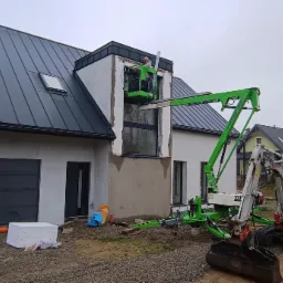 Montaż okna na wysokości w nowym domu jednorodzinnym, pracownicy na podnośniku koszowym, obok minikoparka, teren budowy.