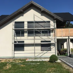 Dom jednorodzinny z zamontowanymi, zewnętrznymi, szarymi żaluzjami fasadowymi, widoczne rusztowanie i drabina oparta o ścianę budynku.