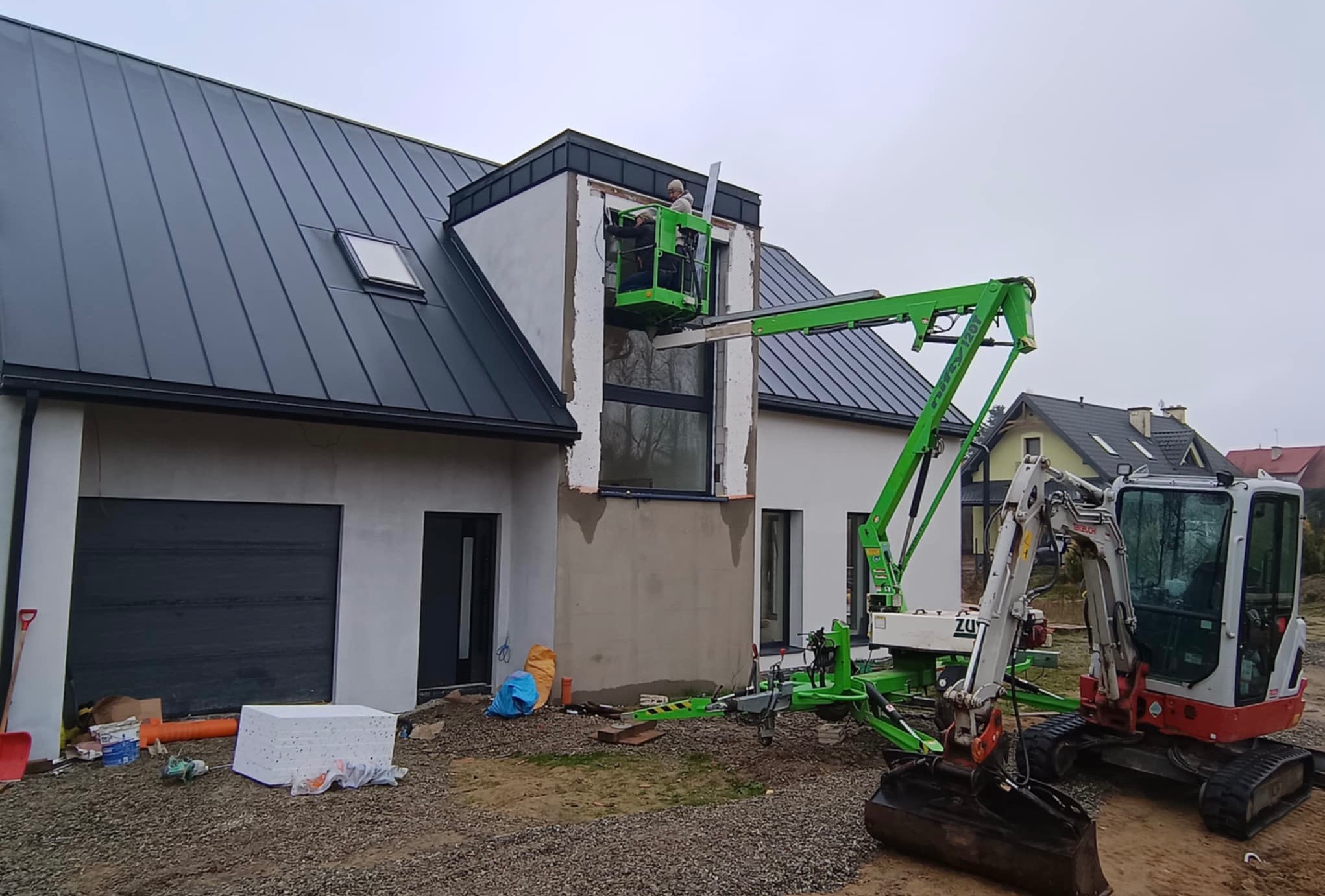 Montaż okna na wysokości w nowym domu jednorodzinnym, pracownicy na podnośniku koszowym, obok minikoparka, teren budowy.