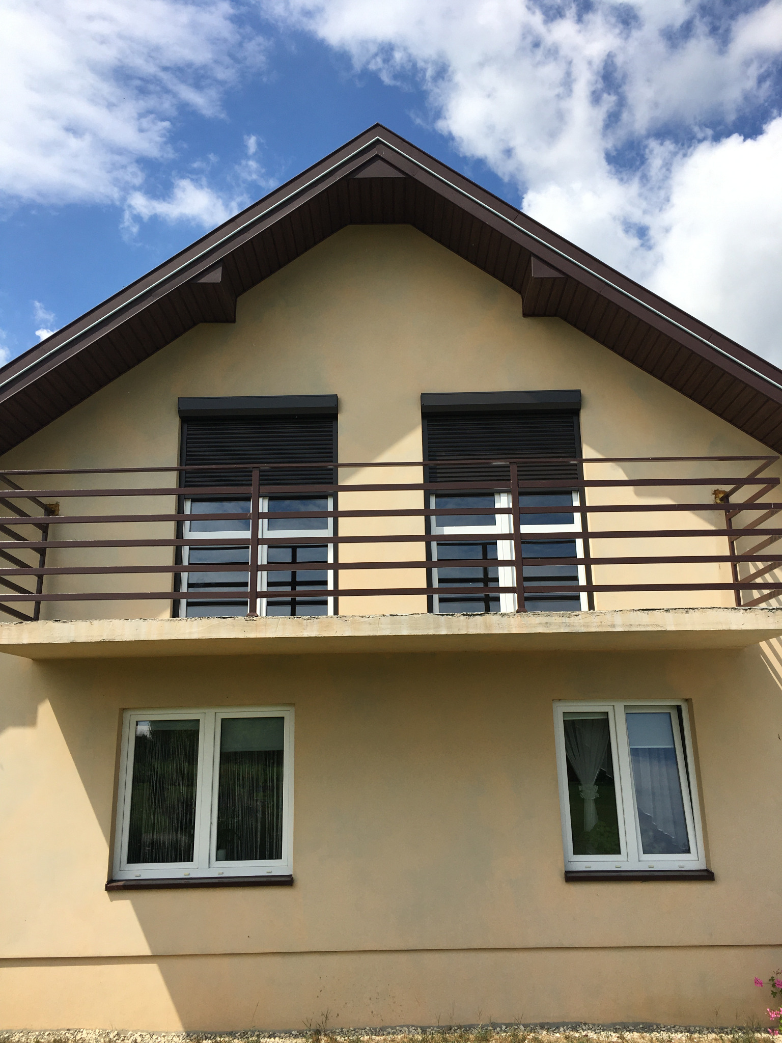 Dwupiętrowy dom z beżową elewacją, brązowym dachem i balkonem z brązową balustradą. Na górnych oknach zamontowane czarne rolety zewnętrzne.