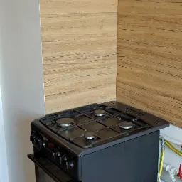 Czarna kuchenka gazowa z piekarnikiem, ustawiona w rogu kuchni z panelami ściennymi imitującymi drewno.