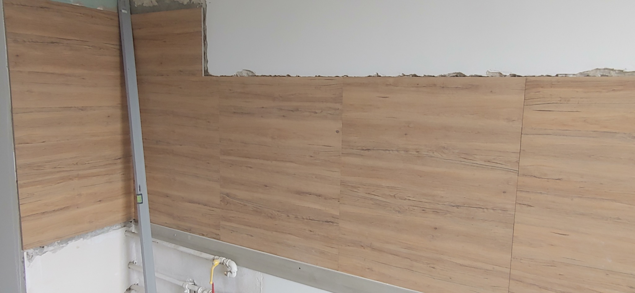 Ściana wyłożona panelami imitującymi drewno, widoczny poziomica i fragmenty surowej ściany w górnej części, instalacja wodna u dołu.