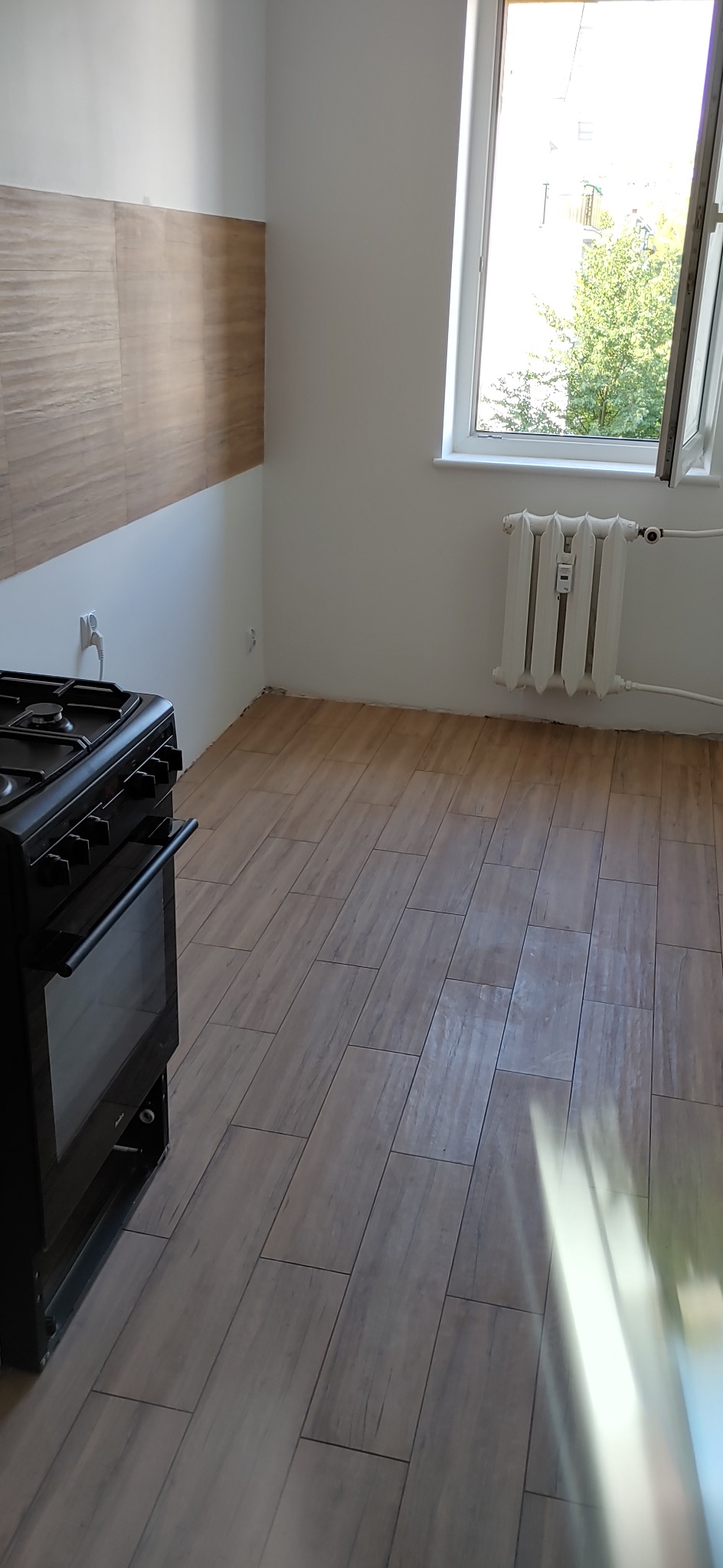 Kuchnia w trakcie prac wykończeniowych: podłoga wyłożona płytkami imitującymi drewno, częściowo wykończona ściana z brązowymi płytkami, białe ściany i okno z widokiem na zieleń, czarny piec.