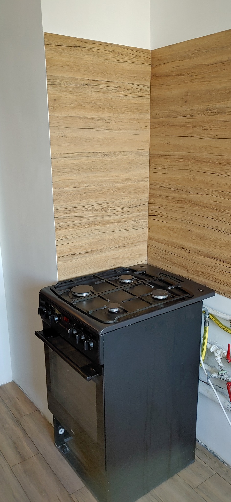 Czarna kuchenka gazowa z piekarnikiem, ustawiona w rogu kuchni z panelami ściennymi imitującymi drewno.