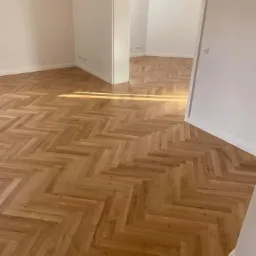 Ułożona w jodełkę podłoga z paneli w jasnym odcieniu drewna, widok na fragment pomieszczenia z białymi ścianami i otwartymi drzwiami do kolejnego pokoju.