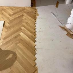 TraczykWykończenia - Fragment podłogi z ułożonym parkietem w jodełkę, widoczny laserowy poziom wyznaczający linię montażu, klej montażowy i białe płyty podkładowe.