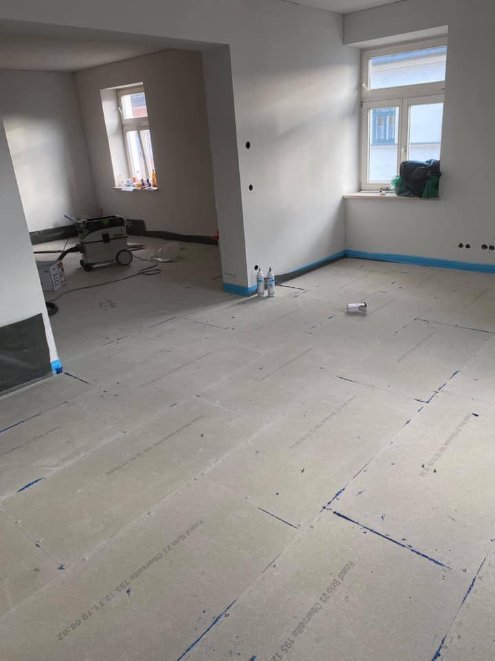 Przygotowanie podłoża pod nową podłogę: ułożone płyty izolacyjne z widocznym nadrukiem producenta, zabezpieczone ściany taśmą malarską, odkurzacz przemysłowy w tle.