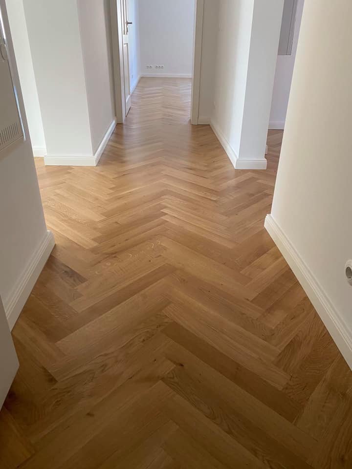 Ułożona parkietowa podłoga w jodełkę w korytarzu z białymi ścianami i listwami przypodłogowymi, widok z perspektywy wejścia.