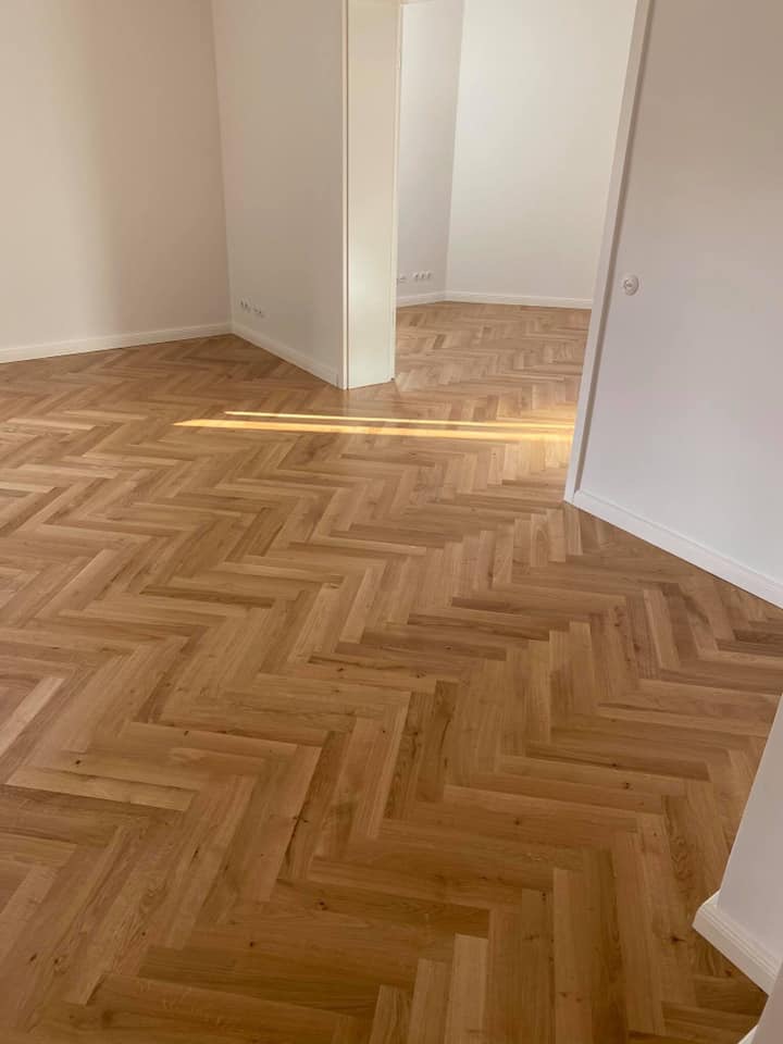 Ułożona w jodełkę podłoga z paneli w jasnym odcieniu drewna, widok na fragment pomieszczenia z białymi ścianami i otwartymi drzwiami do kolejnego pokoju.