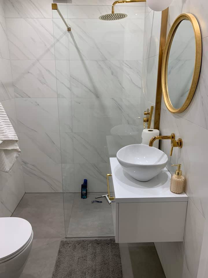 Nowoczesna łazienka z białymi marmurkowymi płytkami, złotą armaturą i okrągłym lustrem, kabina prysznicowa ze szklaną ścianką, biała umywalka nablatowa, widoczne akcesoria łazienkowe i szczoteczka...