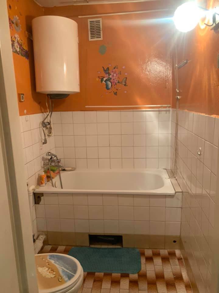 Pomarańczowa łazienka przed remontem: widok na wannę obudowaną białymi płytkami, bojler na ścianie i wzory motyli na pomalowanej ścianie.