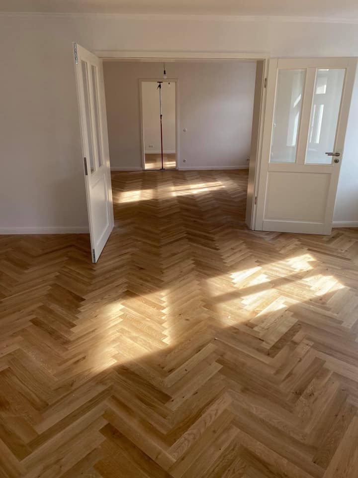 Jasne, drewniane panele ułożone w jodełkę w pustym pokoju z otwartymi, białymi drzwiami dwuskrzydłowymi; widoczne promienie słoneczne.