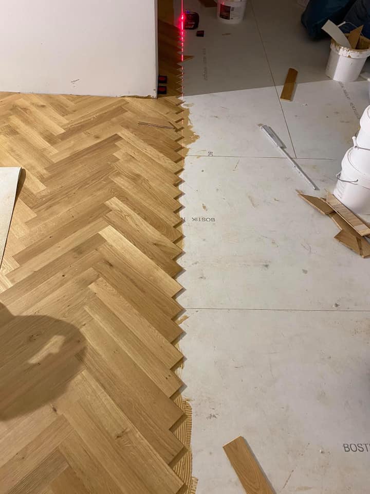 Fragment podłogi z ułożonym parkietem w jodełkę, widoczny laserowy poziom wyznaczający linię montażu, klej montażowy i białe płyty podkładowe.