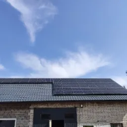 Instalacja paneli słonecznych na dachu budynku gospodarczego w Kielcach, widoczne niebieskie niebo z chmurami w tle.