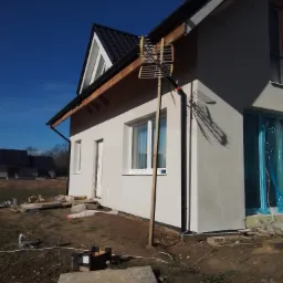 Elewacja domu jednorodzinnego po tynkowaniu, z widoczną anteną opartą o ścianę, okna zabezpieczone folią, teren wokół w trakcie prac porządkowych.