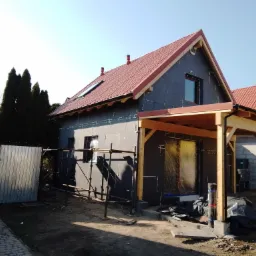 Dom jednorodzinny w trakcie ocieplania, widoczne czarne płyty izolacyjne na elewacji, rusztowania, drewniana konstrukcja zadaszenia ganku, czerwona dachówka i metalowa brama w tle.