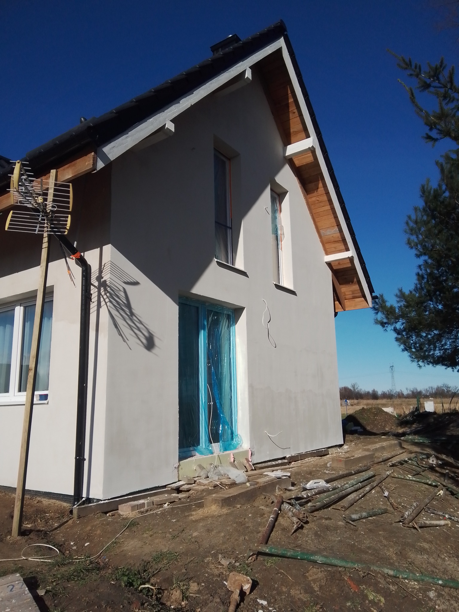 Świeżo otynkowana elewacja domu jednorodzinnego w trakcie prac wykończeniowych, zabezpieczone okna folią, widoczne elementy rusztowania i antena na ścianie bocznej.