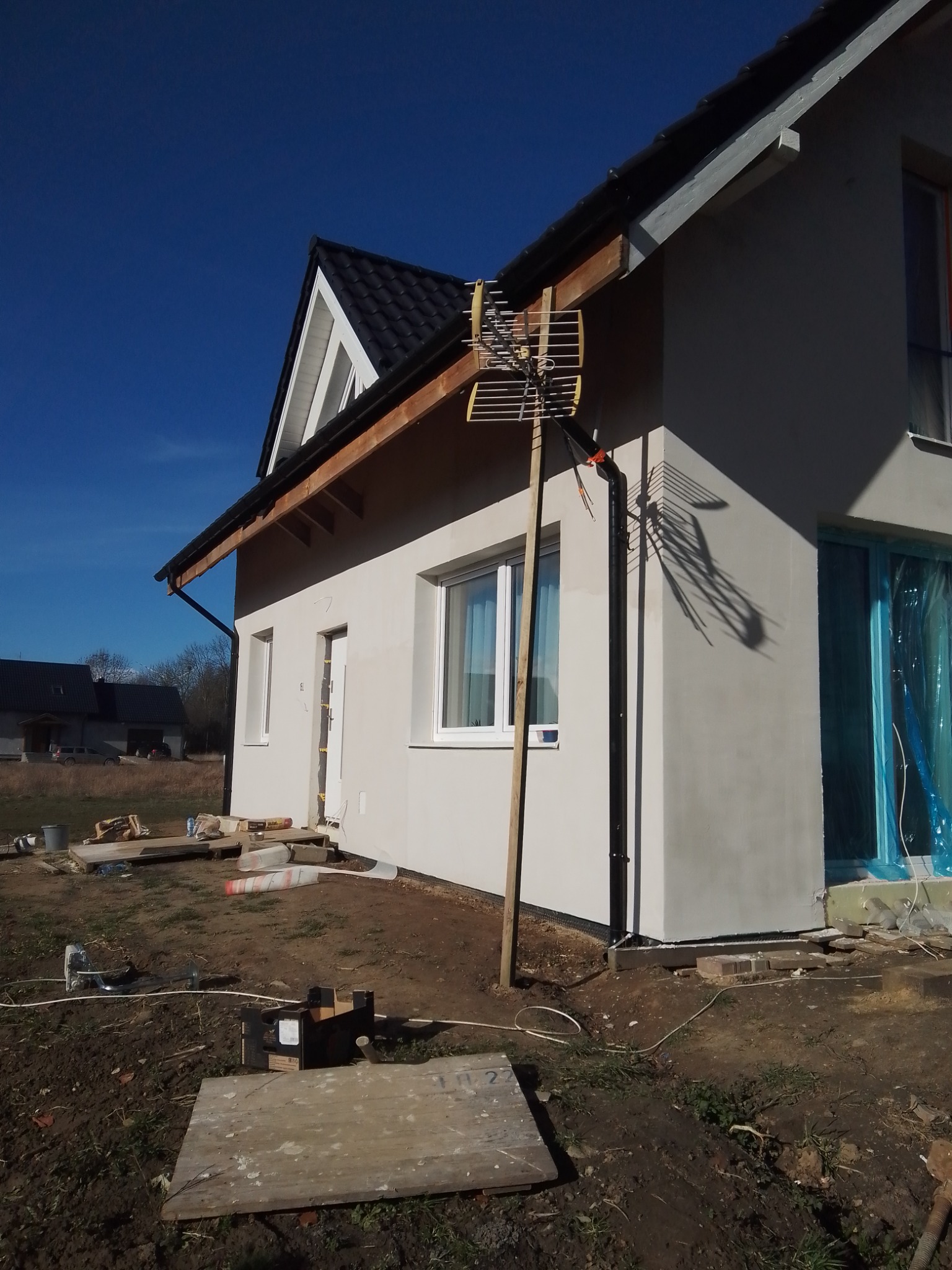 Elewacja domu jednorodzinnego po tynkowaniu, z widoczną anteną opartą o ścianę, okna zabezpieczone folią, teren wokół w trakcie prac porządkowych.