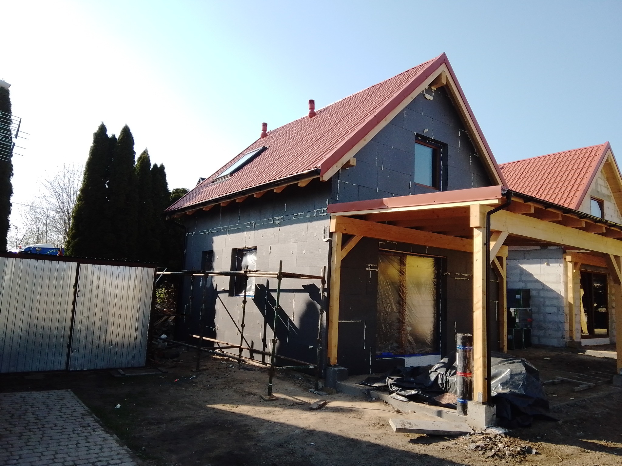 Dom jednorodzinny w trakcie ocieplania, widoczne czarne płyty izolacyjne na elewacji, rusztowania, drewniana konstrukcja zadaszenia ganku, czerwona dachówka i metalowa brama w tle.