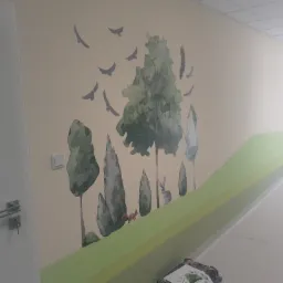 Ściana w korytarzu z ręcznie malowanym motywem leśnym: drzewa, ptaki, wiewiórka i królik na zielonym tle. Częściowo widoczny sprzęt malarski na podłodze.