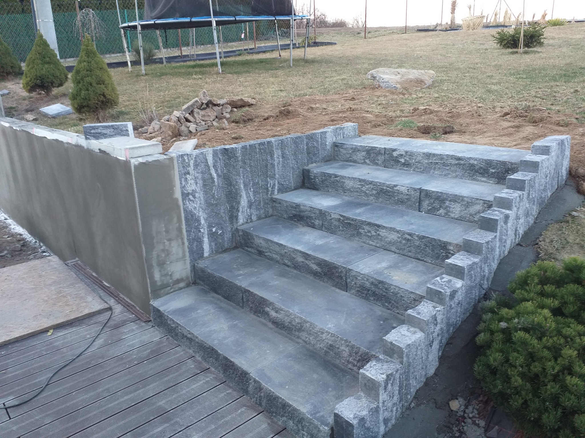 Schody zewnętrzne z kostki granitowej, prowadzące z drewnianego tarasu na trawnik, obok świeżo wzniesiona ściana oporowa z betonu.