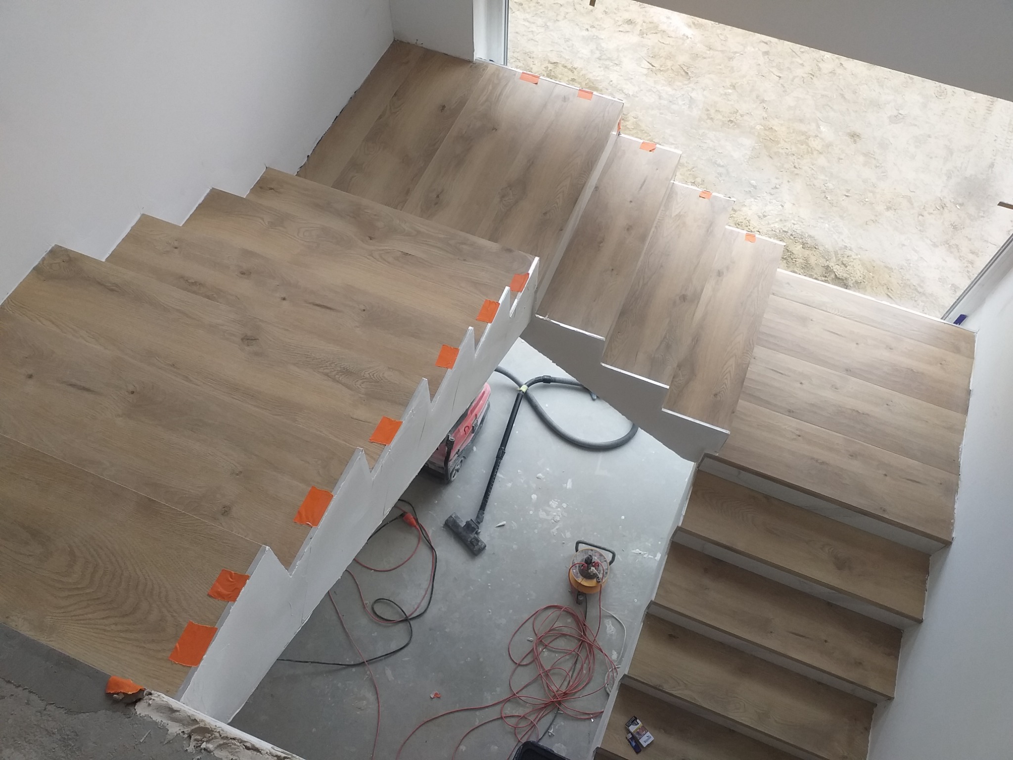 Montaż drewnopodobnych paneli na schodach w trakcie prac wykończeniowych, widoczne narzędzia i zabezpieczenia krawędzi pomarańczową taśmą.