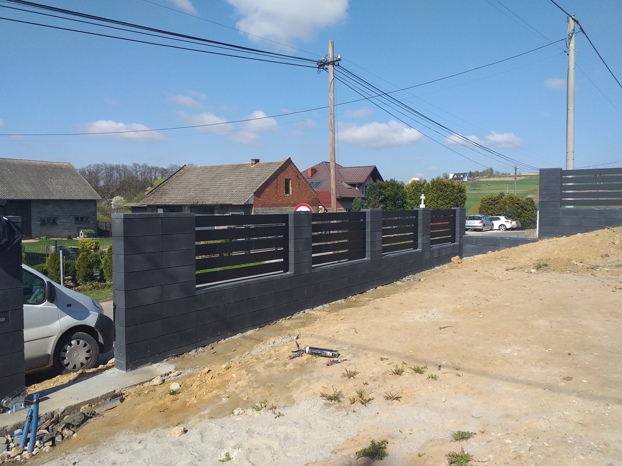 Nowoczesne ogrodzenie z szarych, betonowych bloczków z poziomymi, czarnymi przęsłami, widoczne narzędzia montażowe i samochód dostawczy w tle.