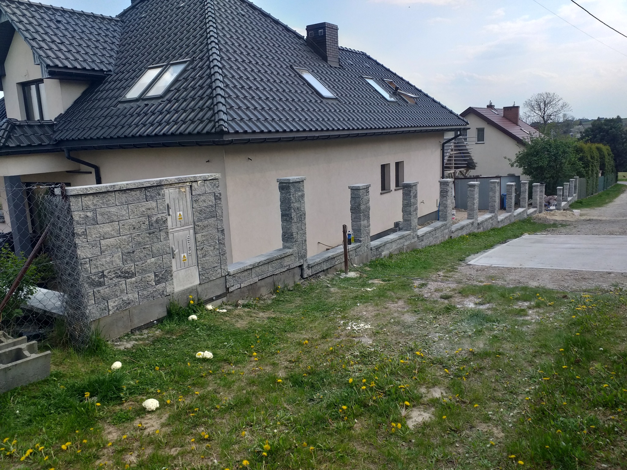 Nowoczesny dom jednorodzinny z szarym dachem i jasną elewacją. Wzdłuż posesji wzniesiony niski, murowany płot z szarych bloczków z elementami dekoracyjnymi w postaci słupków. Widoczne świeże ślady...