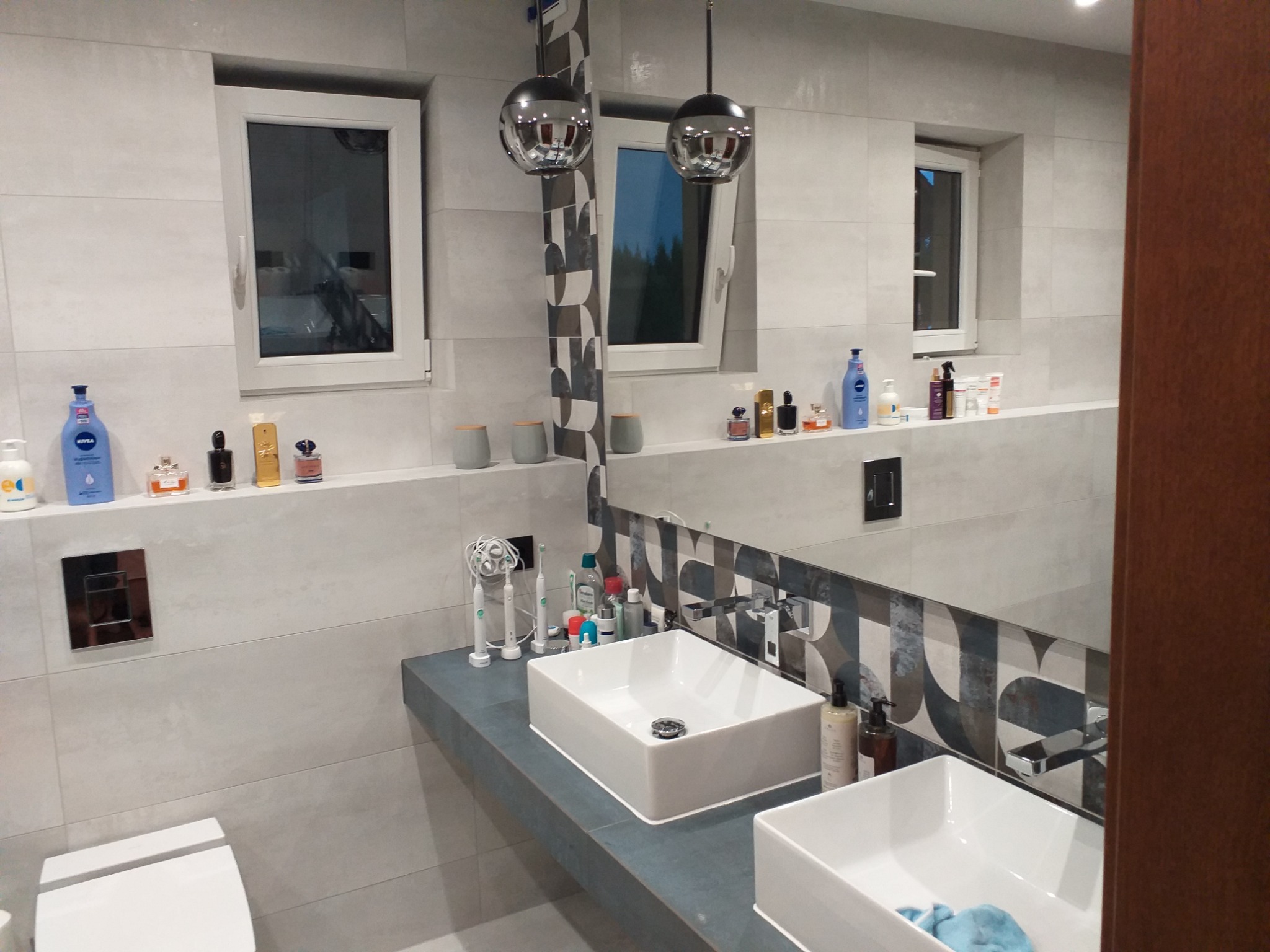 Nowoczesna łazienka z dwoma białymi, prostokątnymi umywalkami nablatowymi na szarym blacie, podwieszane lampy kuliste, jasne płytki imitujące beton, okna z widokiem na zieleń, oraz półki...