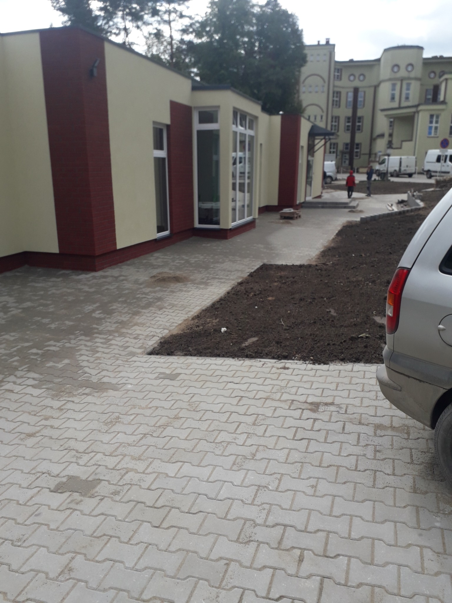 Szara kostka brukowa ułożona w zygzakowaty wzór przed budynkiem z beżową elewacją i ceglanymi wstawkami, obok przygotowane rabaty ziemi i fragment zaparkowanego samochodu.