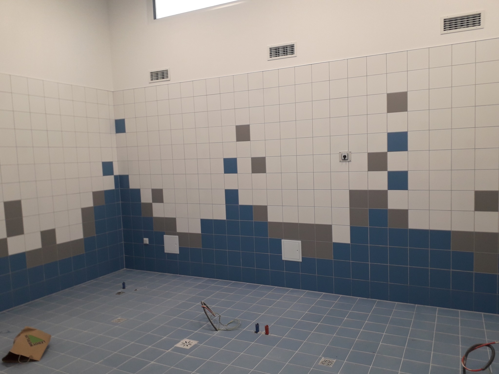 Wnętrze łazienki w trakcie remontu z niebiesko-szaro-białą mozaiką z płytek na ścianach, niebieskie płytki podłogowe, widoczne narzędzia i torba budowlana Leroy Merlin.