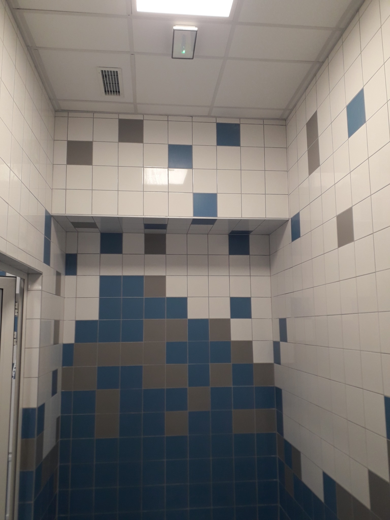 Ściany łazienki wyłożone białymi, szarymi i niebieskimi kwadratowymi płytkami ceramicznymi, tworzącymi wzór. Widoczna półka również wyłożona płytkami, sufit podwieszany z kratką wentylacyjną...