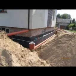 Odsłonięte fundamenty domu z izolacją pionową, widoczne rury drenarskie i obsypka piaskiem, w tle budynek gospodarczy.