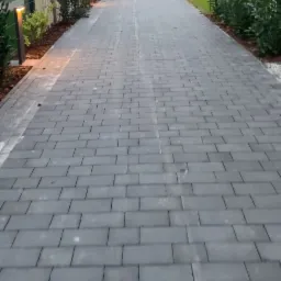 Długa, szara droga z kostki brukowej prowadząca do nowoczesnego domu w Berlinie, otoczona zielenią i oświetlona lampami ogrodowymi.