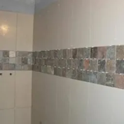 Ściana łazienki wyłożona jasnymi, beżowymi płytkami ceramicznymi z poziomym pasem dekoracyjnym z mozaiki w odcieniach szarości, brązu i beżu.