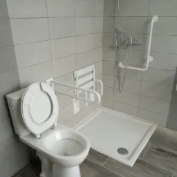 Jasna łazienka z udogodnieniami dla osób niepełnosprawnych: toaleta z uchylnymi poręczami, składane siedzisko prysznicowe i brodzik z niskim progiem.