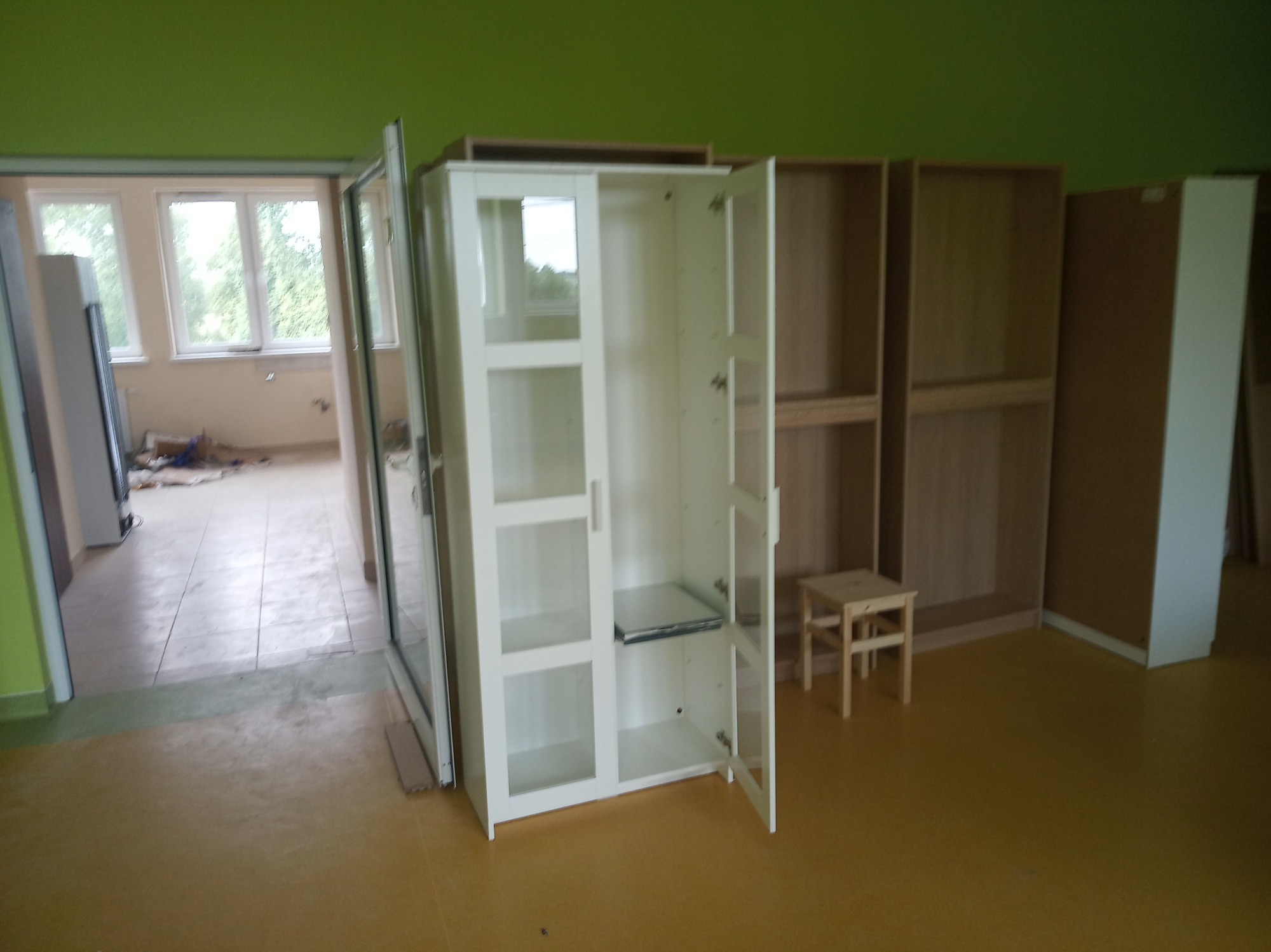 Biała szafa z przeszklonymi drzwiami, otwarte skrzydło, widoczne półki, w tle niedokończone zabudowy meblowe i mały stołek, zielona ściana, pomieszczenie w trakcie prac wykończeniowych.