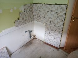 Fragment ściany w trakcie układania mozaiki w narożniku łazienki, widoczne niedokończone prace, fragment instalacji wodnej i surowa podłoga.