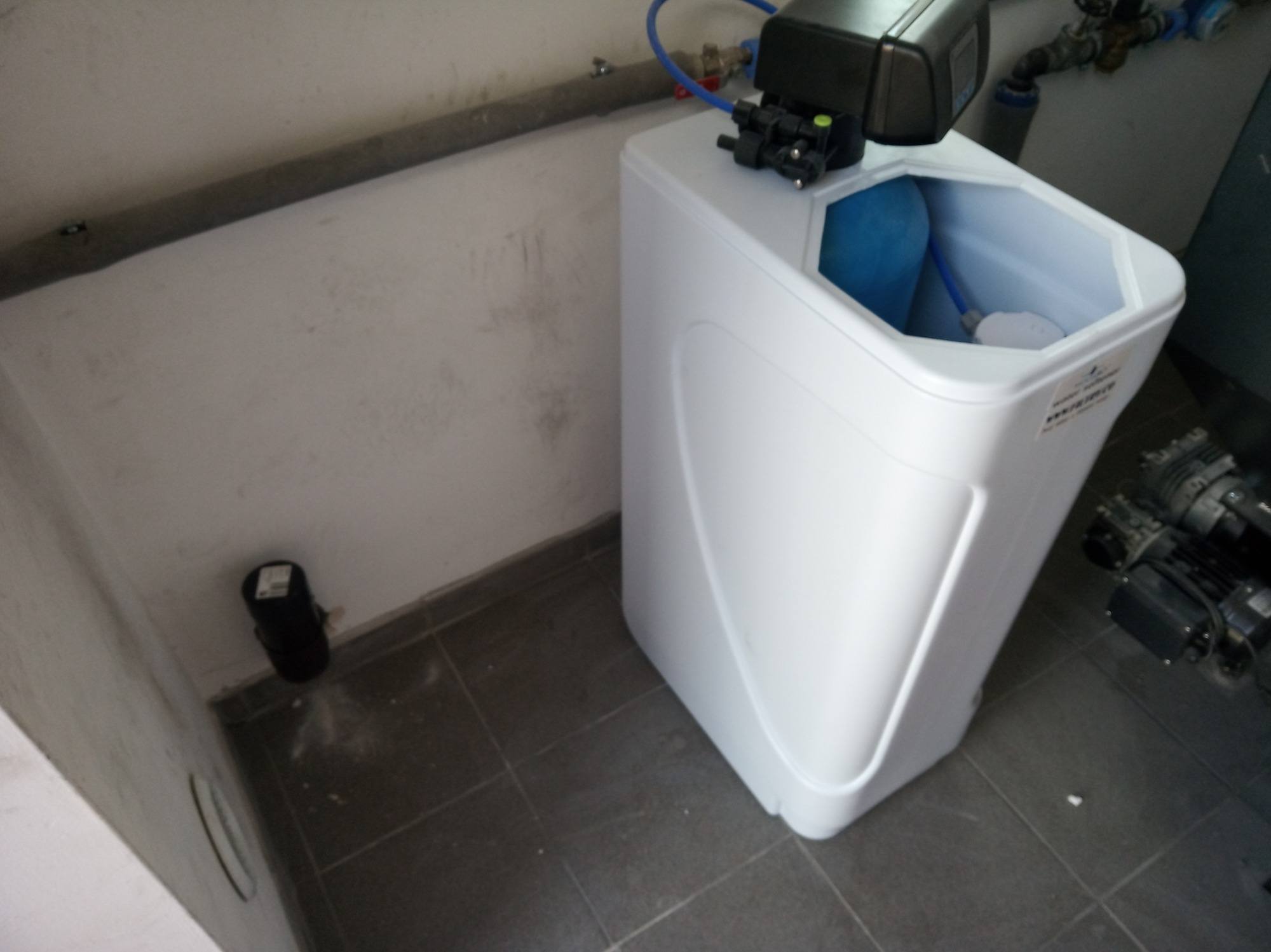 Biały zmiękczacz wody z niebieskimi rurkami podłączony do instalacji, widoczny na tle szarej podłogi i białej ściany w pomieszczeniu technicznym.