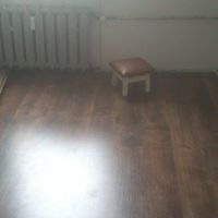 Błyszcząca, ciemnobrązowa podłoga z paneli, mały biały stołek z brązowym siedziskiem, biały grzejnik żeliwny na ścianie, słabe oświetlenie.